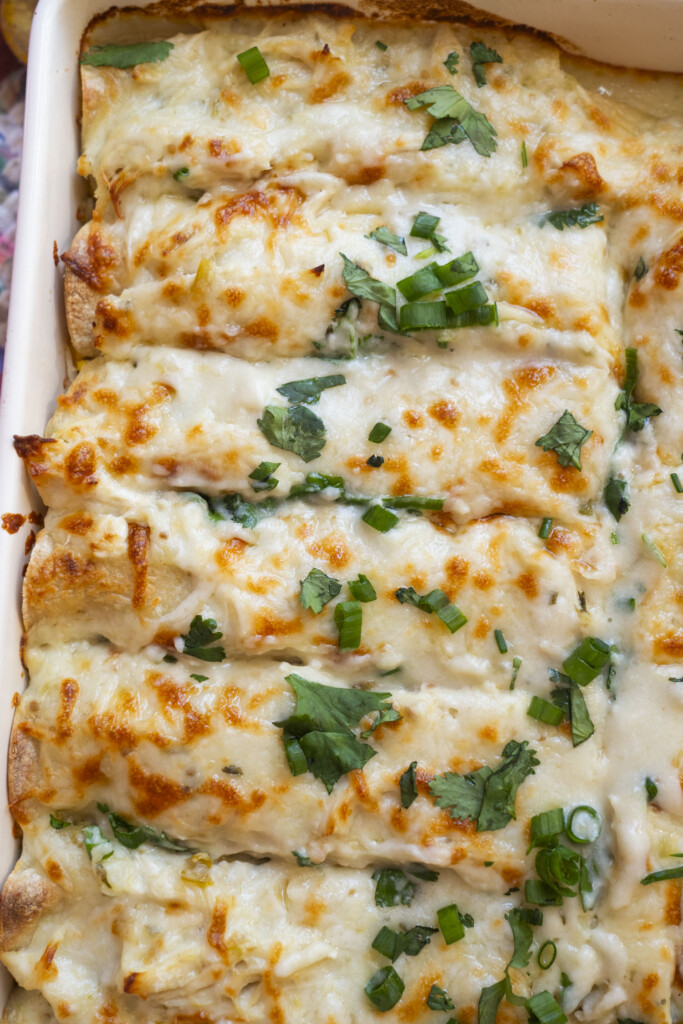 A close-up of baked enchiladas topped with melted cheese, chopped green onions, and cilantro in a rectangular dish. The enchiladas are golden brown and neatly arranged side by side.