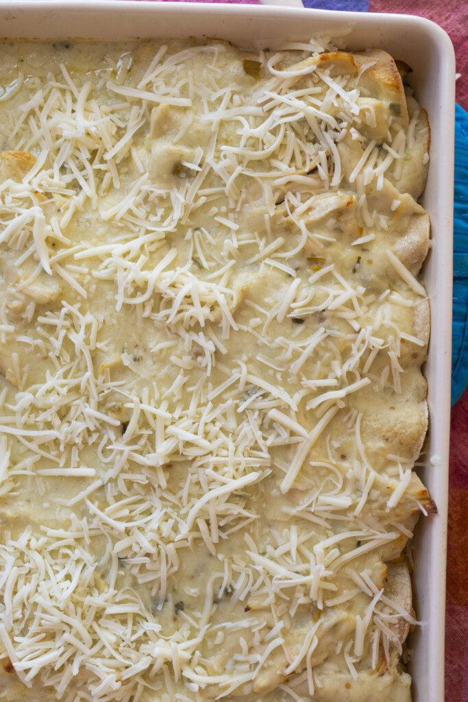 A close-up of a casserole dish filled with enchiladas topped with creamy sauce and shredded cheese, ready to be baked.