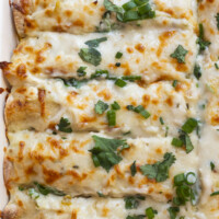 Golden, cheesy enchiladas topped with melted cheese, chopped green onions, and fresh cilantro, arranged closely together in a baking dish.