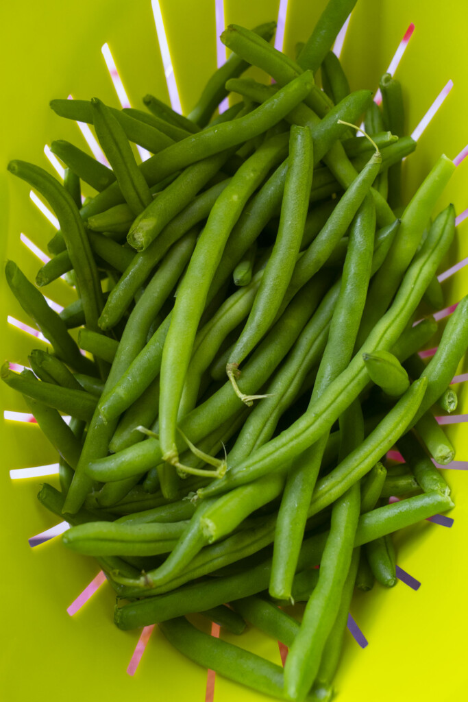 A pile of fresh green beans sits in a bright yellow colander with pink-tipped handles visible around the edges.