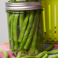 A glass mason jar filled with fresh green beans, packed vertically in a clear pickling liquid, sits on a colorful surface with a few loose green beans nearby.