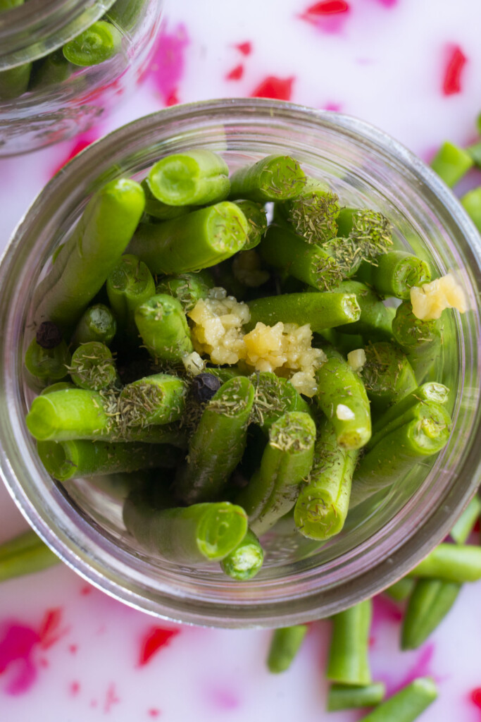 Close-up of a glass jar filled with fresh green bean pieces, minced garlic, and dried dill, with some beans and pink pieces scattered on a white surface in the background.