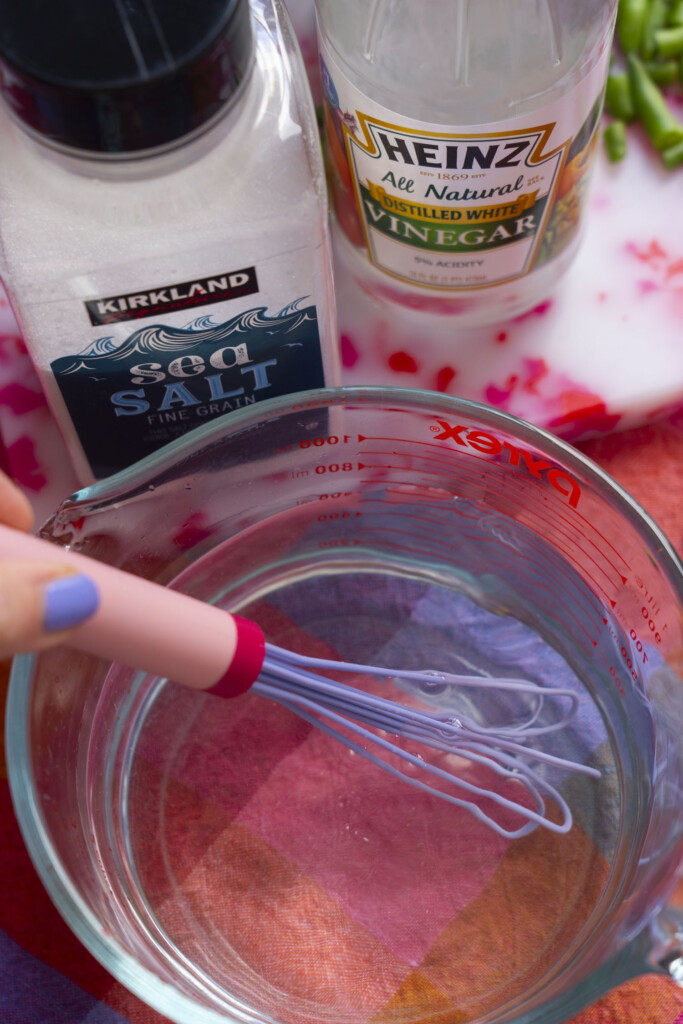 A hand holding a whisk stirs clear liquid in a glass measuring cup. Nearby are containers of Kirkland sea salt and Heinz distilled white vinegar on a colorful surface.
