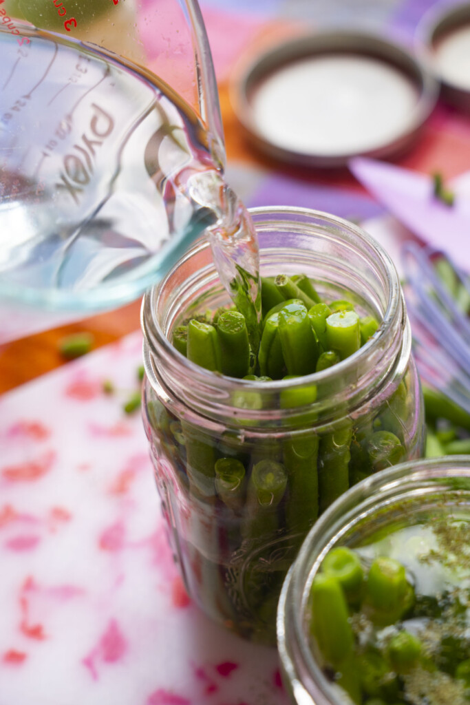 A close-up of water being poured from a glass measuring cup into a mason jar filled with fresh green beans, on a colorful kitchen surface with blurred jars and utensils in the background.