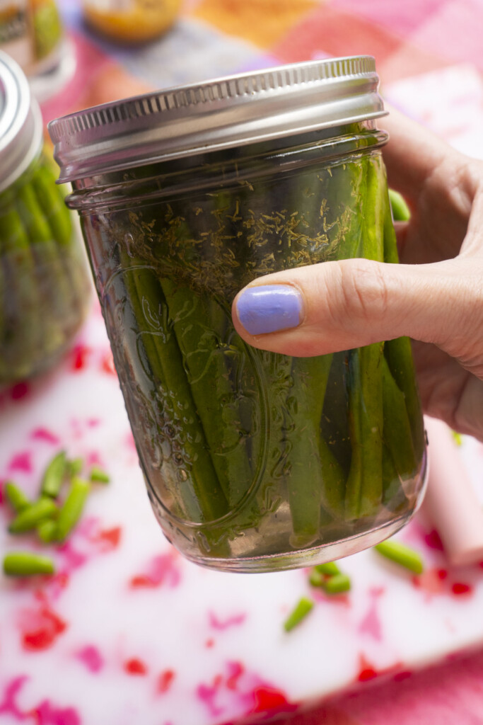 A hand with light purple nail polish holds a mason jar filled with pickled green beans. The background shows a blurred cutting board with green bean pieces and colorful fabric.