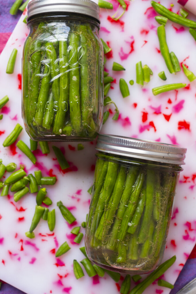 Two mason jars filled with green beans are placed on a white and pink surface, with chopped green beans scattered around them. The jars have metal lids and are arranged at an angle.
