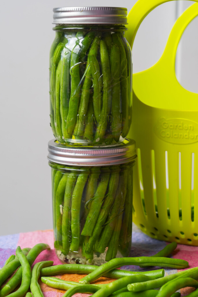 Two jars filled with pickled green beans are stacked on top of each other, next to a bright yellow colander and several loose green beans on a colorful cloth.