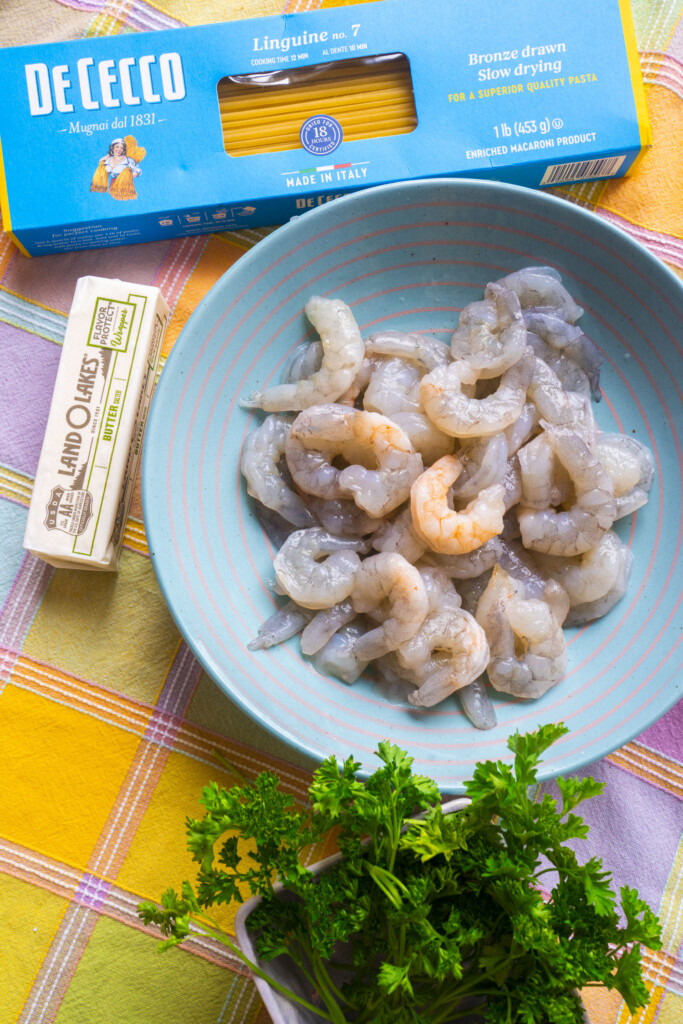 A plate of raw shrimp, a stick of butter, a box of De Cecco linguine pasta, and fresh parsley are arranged on a yellow plaid tablecloth.