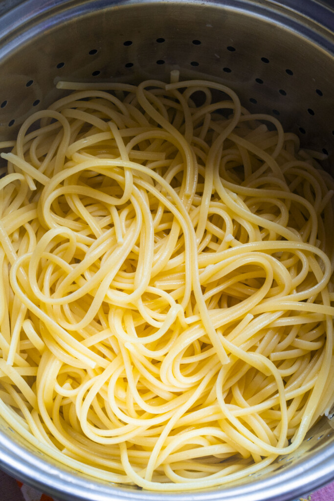 Cooked spaghetti noodles in a metal colander, with steam holes visible in the background. The noodles appear freshly boiled and slightly glossy.