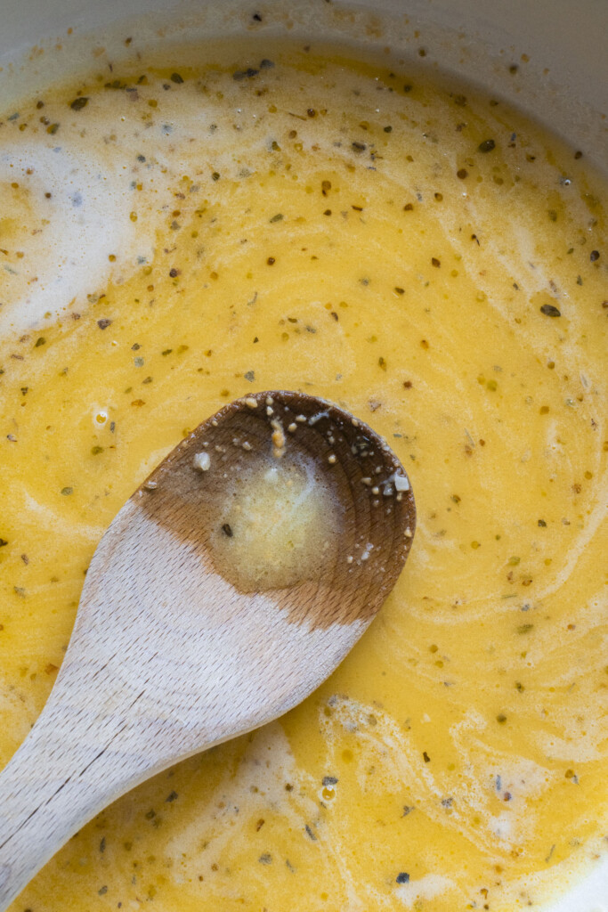 A close-up of a wooden spoon resting on a mixture of whisked eggs and seasonings in a bowl, showing swirls of yellow and specks of herbs.