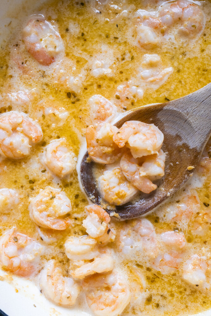 Cooked shrimp in a creamy, orange-colored garlic butter sauce, being stirred with a wooden spoon in a pan. The shrimp are pink and coated in the rich, bubbly sauce.