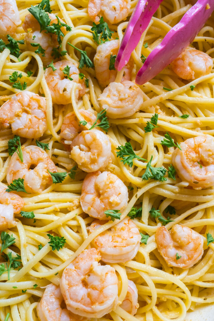 Linguine pasta topped with cooked shrimp and garnished with chopped parsley, with pink serving tongs resting on top.
