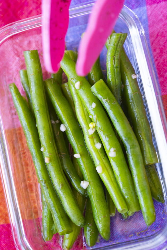 A glass container filled with pickled green beans sprinkled with minced garlic. Pink tongs are seen at the top, holding one of the green beans. The background is a colorful, patterned cloth.