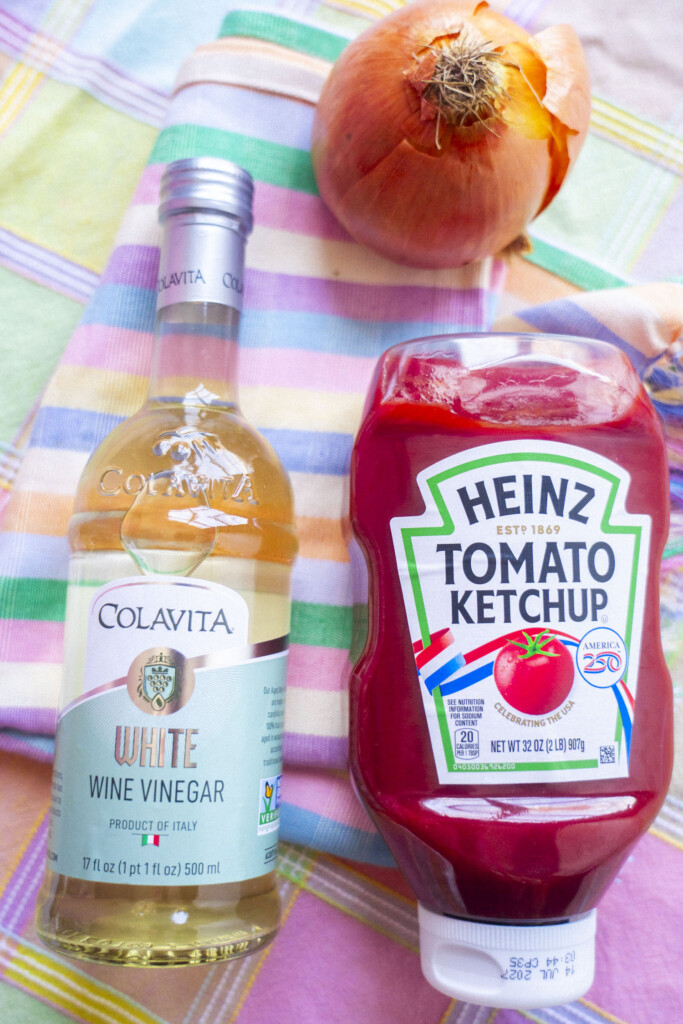 A bottle of Colavita white wine vinegar, a bottle of Heinz tomato ketchup, and a whole onion are placed on a pastel plaid cloth.