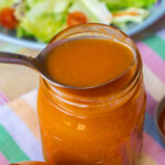 A metal spoon scoops orange salad dressing from a glass jar, with a blurred plate of salad and colorful tablecloth in the background.