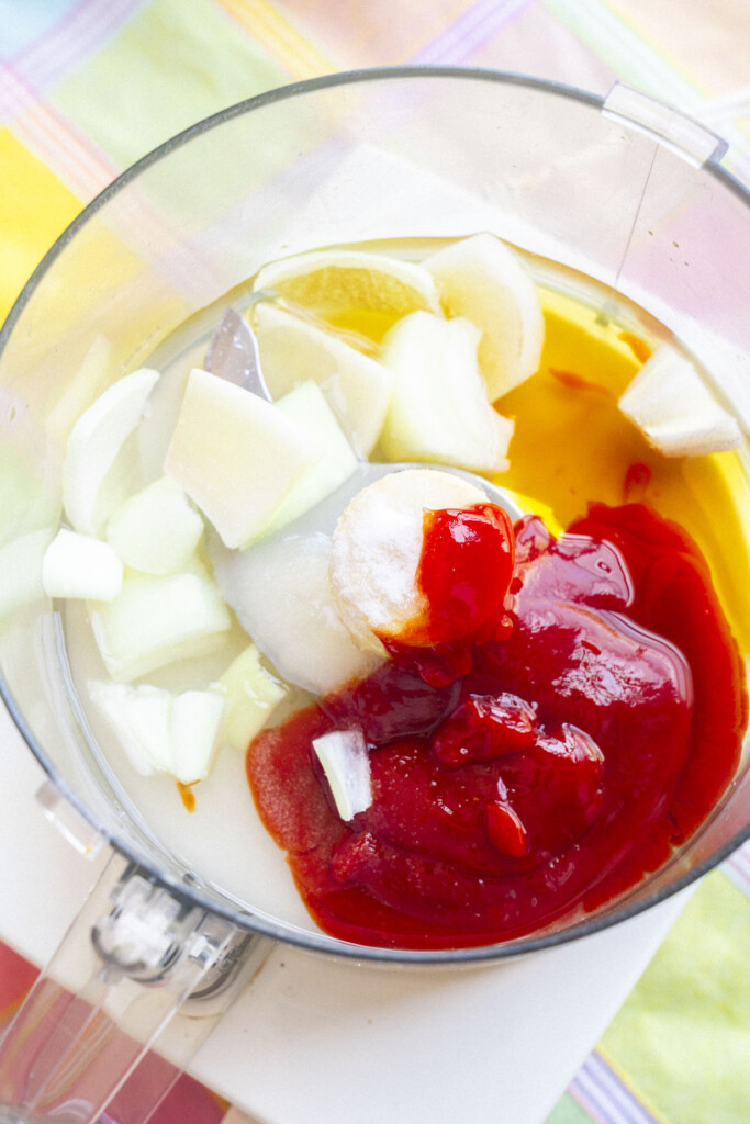 A food processor bowl containing chopped onions, ketchup, vegetable oil, vinegar, lemon wedge, and sugar, ready to be blended, sits on a colorful striped surface.
