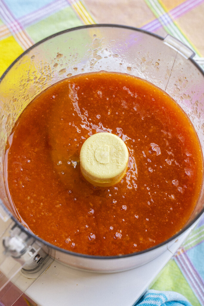 A food processor bowl filled with a smooth, bright red-orange sauce, sitting on a colorful plaid tablecloth.