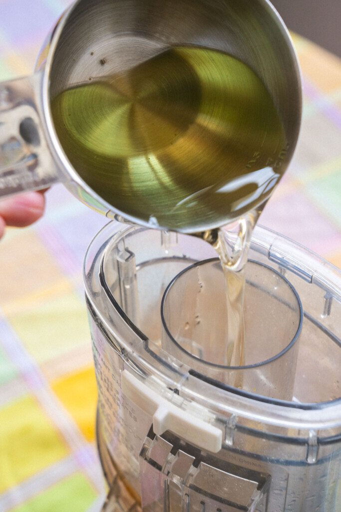 A hand pours olive oil from a metal measuring cup into the spout of a food processor on a colorful, checkered surface.