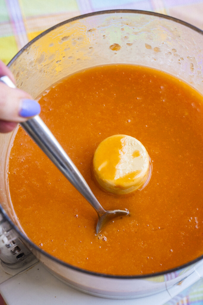 A close-up of a food processor bowl with an orange salad dressing inside. A hand with purple nail polish holds a spoon stirring the mixture. The food processor blade is partially visible in the center.
