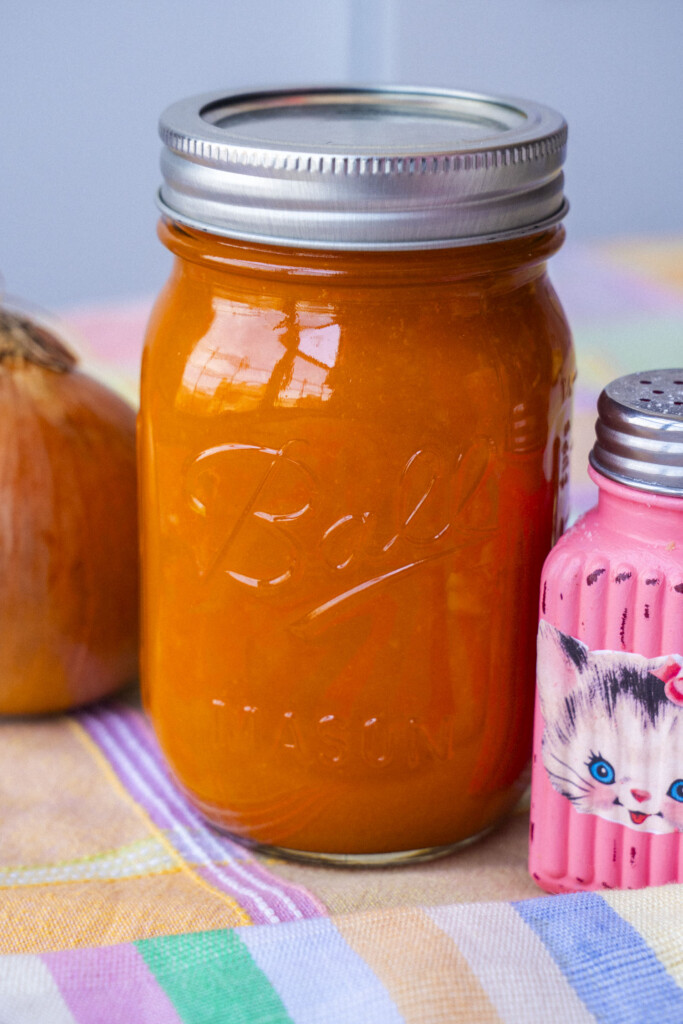 A glass mason jar filled with orange salad dressing sits on a colorful cloth, next to an onion and a pink salt shaker decorated with a cat illustration.