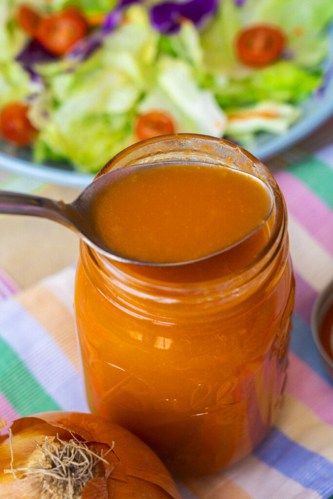 A spoonful of salad dressing is held above a mason jar filled with the same dressing. In the background, there is a fresh salad and part of an onion. The scene is set on a colorful cloth.