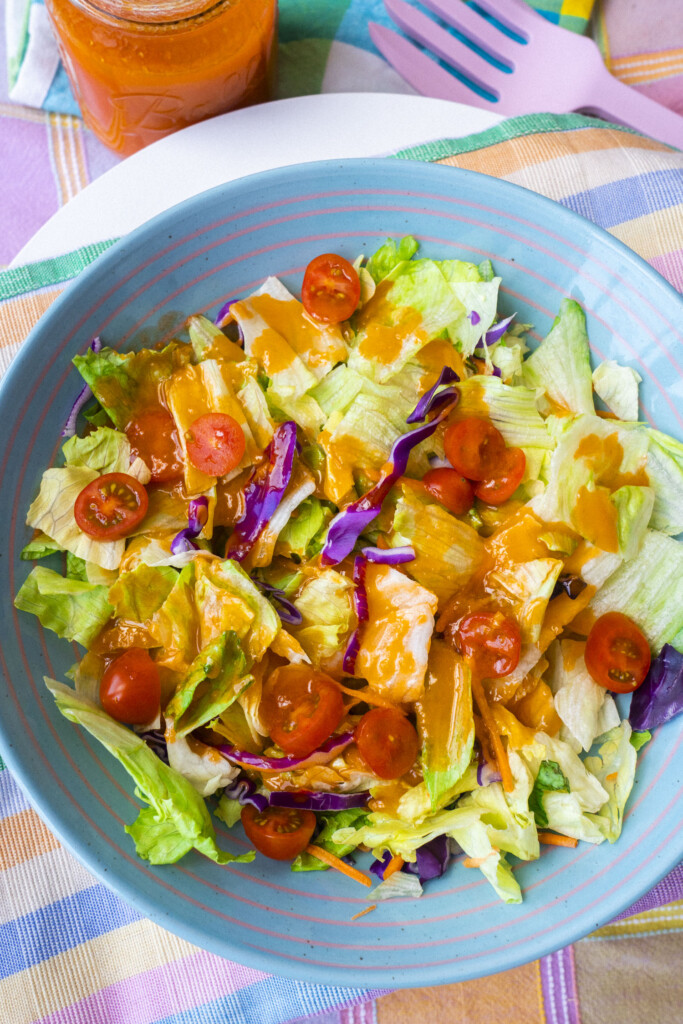 A blue bowl filled with salad made of lettuce, cherry tomatoes, and purple cabbage, topped with orange dressing. A jar of dressing, a pink fork, and a colorful plaid napkin are beside the bowl.