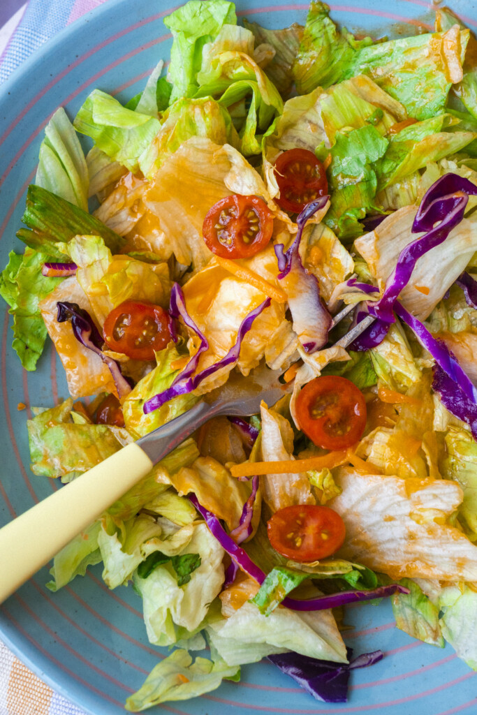A colorful salad with chopped lettuce, cherry tomatoes, shredded purple cabbage, and orange dressing is served on a blue plate with a yellow fork.