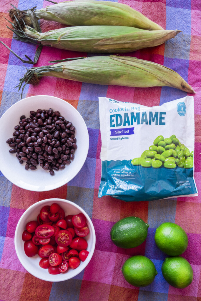 Three ears of corn, a bowl of black beans, a bowl of halved cherry tomatoes, a bag of edamame, and three limes on a colorful plaid tablecloth.
