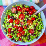 A colorful salad with cherry tomatoes, black beans, corn, and edamame in a blue bowl, with a fork on the side and a multicolored cloth beneath.