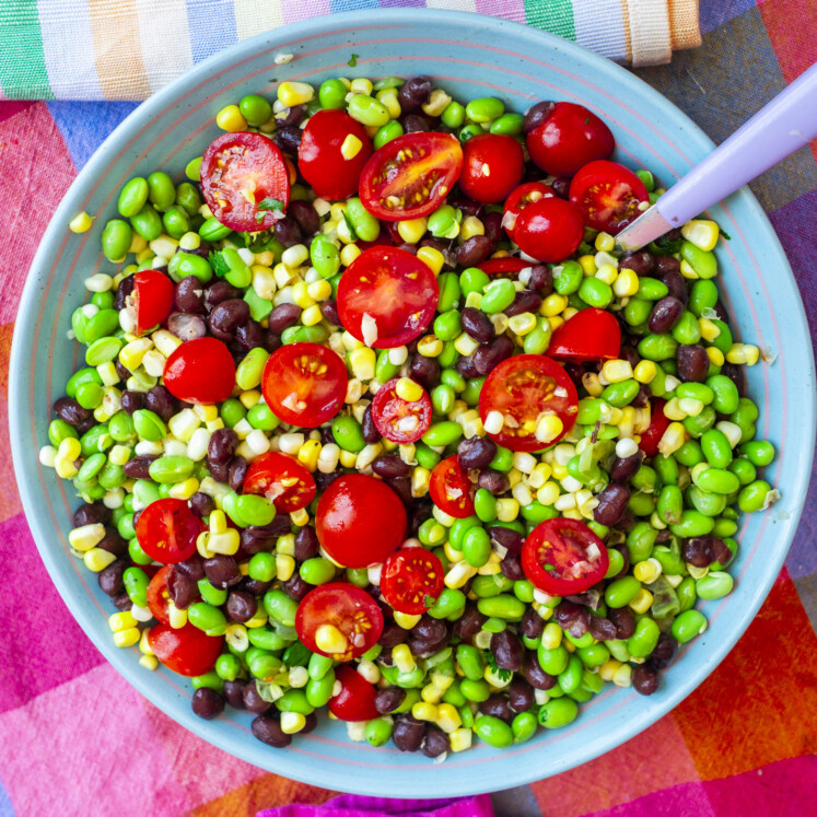 A colorful salad with cherry tomatoes, black beans, corn, and edamame in a blue bowl, with a fork on the side and a multicolored cloth beneath.