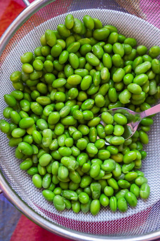 Bright green edamame beans rest in a metal mesh strainer, with a silver spoon partially visible on the right side. The background shows hints of a colorful surface or cloth.