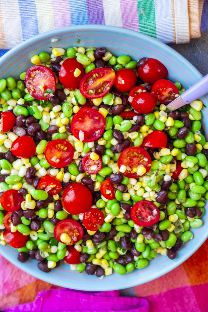 A colorful salad with cherry tomatoes, black beans, edamame, and corn in a blue bowl, with a spoon and striped napkin beside it.
