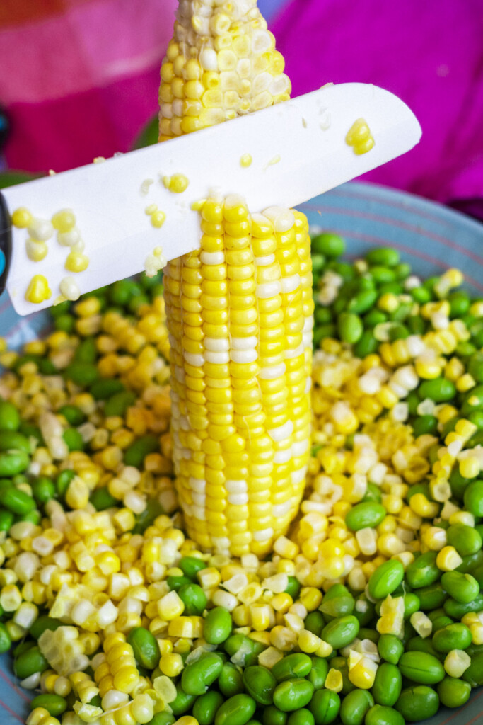 A knife is slicing kernels off a vertical ear of corn, with corn kernels and green edamame beans scattered on a plate beneath.