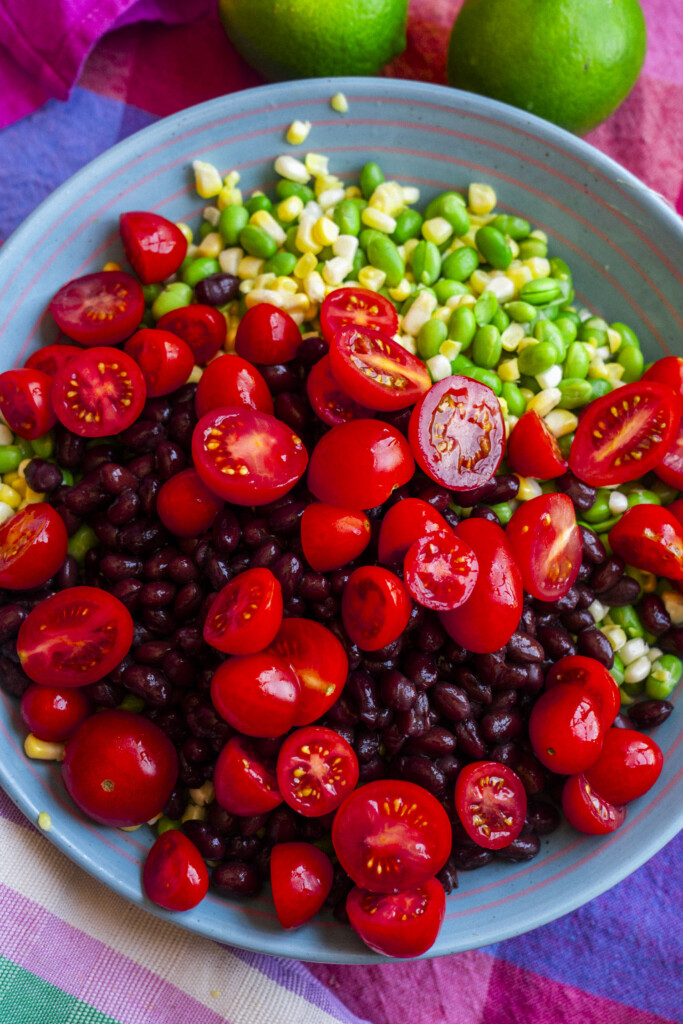 A bowl filled with black beans, halved cherry tomatoes, corn, and edamame, set on a colorful tablecloth with whole limes visible in the background.