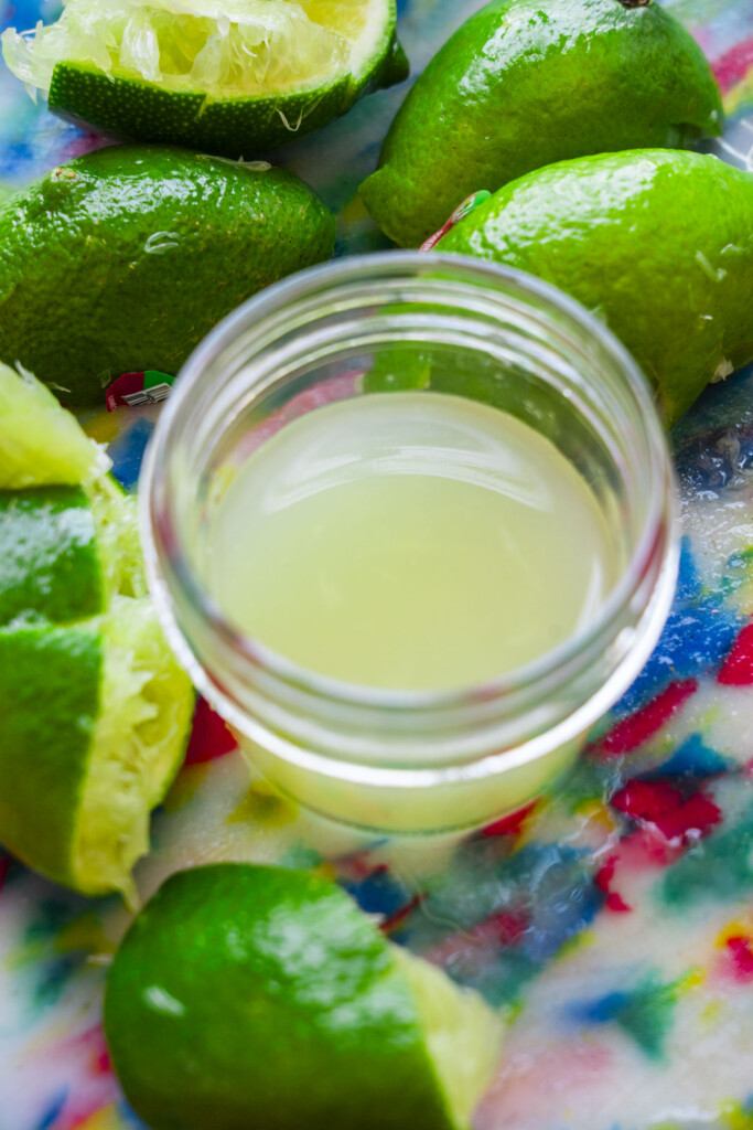 A small glass jar of lime juice surrounded by freshly squeezed lime halves on a colorful, patterned surface.