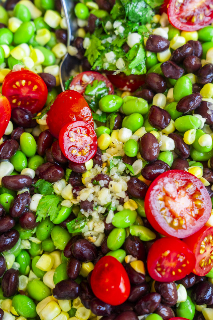 Close-up of a colorful salad with black beans, edamame, corn, halved cherry tomatoes, chopped cilantro, and a spoon visible in the background.