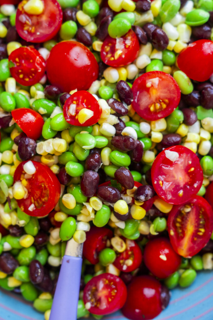 Colorful salad with cherry tomatoes, black beans, edamame, corn, and green peas, all mixed together. A purple spoon is partially visible in the lower left corner of the image.