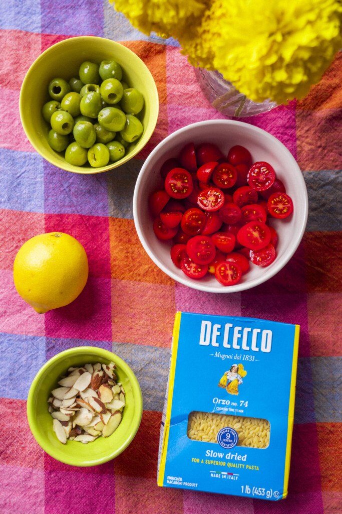 A flat lay of green olives, halved cherry tomatoes, a lemon, sliced almonds with green grapes, and a De Cecco orzo pasta box on a colorful checkered cloth with yellow flowers visible in the corner.