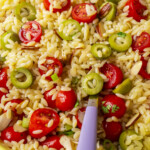 Close-up of orzo pasta salad with sliced green olives, halved cherry tomatoes, chopped green onions, and slivered almonds. A purple-handled fork is partially visible among the colorful ingredients.