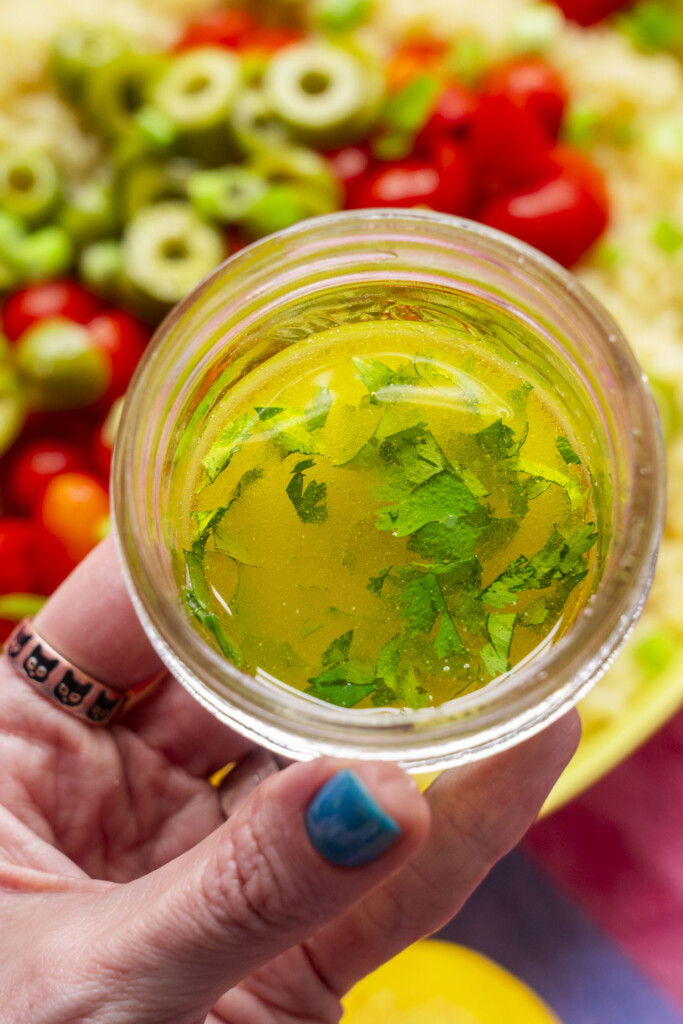 A hand with blue nail polish holds a small jar of yellow salad dressing with chopped herbs, in front of a colorful salad with tomatoes and sliced green olives.