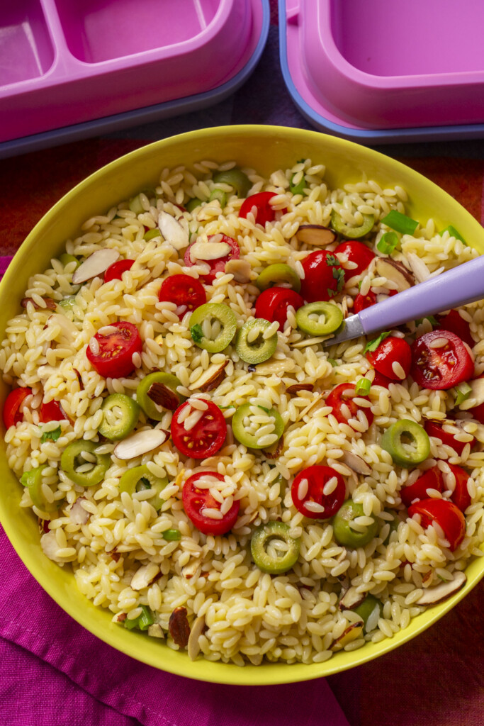 A yellow bowl filled with orzo pasta, halved cherry tomatoes, sliced green olives, and almond slivers, with a fork resting inside. Pink lunch boxes are visible in the background.