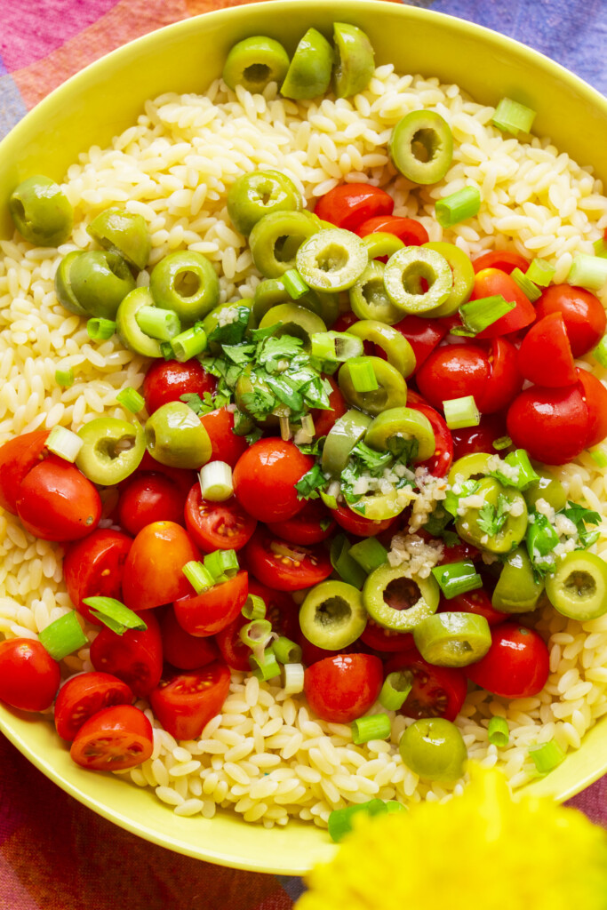 A bowl of orzo pasta topped with sliced green olives, halved cherry tomatoes, chopped green onions, and fresh herbs, set on a colorful tablecloth.