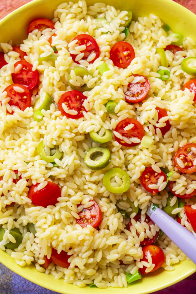 A bowl of orzo pasta salad with halved cherry tomatoes, sliced green olives, and green onions, mixed together with a light dressing. A white serving spoon is partially visible in the bowl.