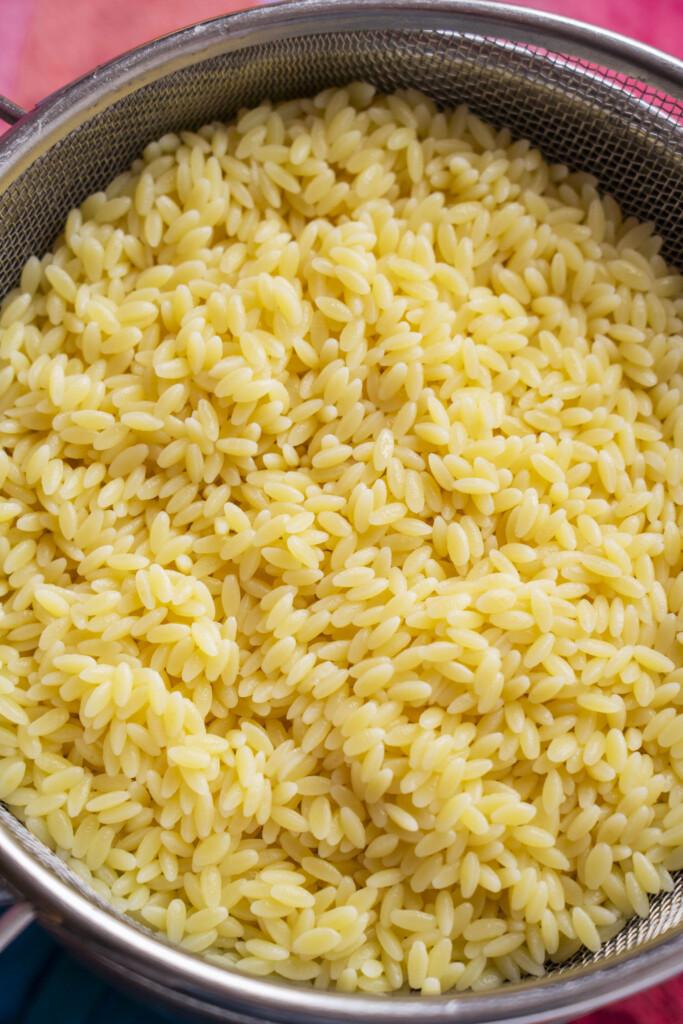 Cooked orzo pasta is draining in a metal mesh strainer. The small, yellow, rice-shaped pasta pieces fill the strainer, and hints of a colorful surface can be seen underneath.