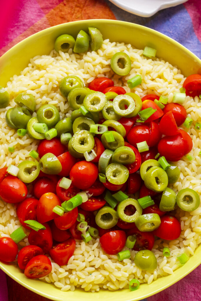 A bowl of orzo pasta topped with halved cherry tomatoes, sliced green olives, and chopped green onions sits on a colorful tablecloth.