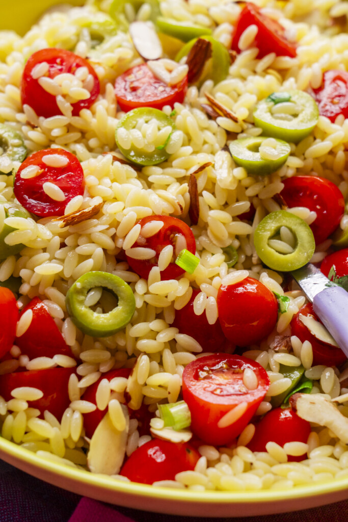 Close-up of orzo pasta salad with halved cherry tomatoes, sliced green olives, scallions, and almond slivers, served in a yellow bowl with a purple-handled fork.