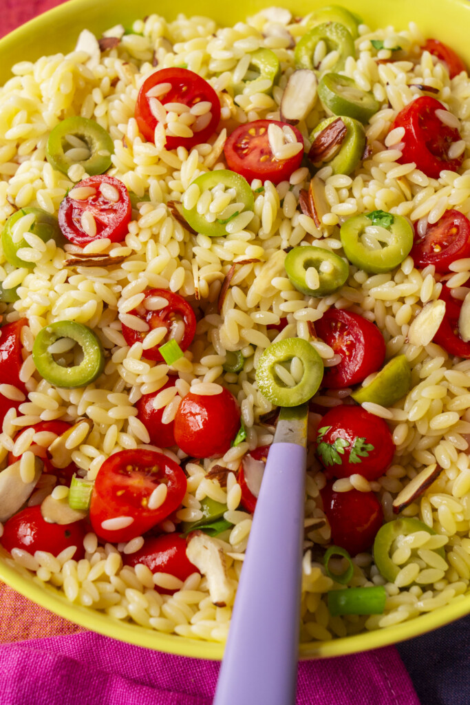A bowl of orzo pasta salad with sliced green olives, cherry tomatoes, slivered almonds, and green onions, with a purple-handled fork resting inside.