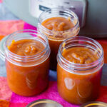 Three glass jars filled with thick, amber-colored apple butter sitting on a checkered cloth in front of a slow cooker.