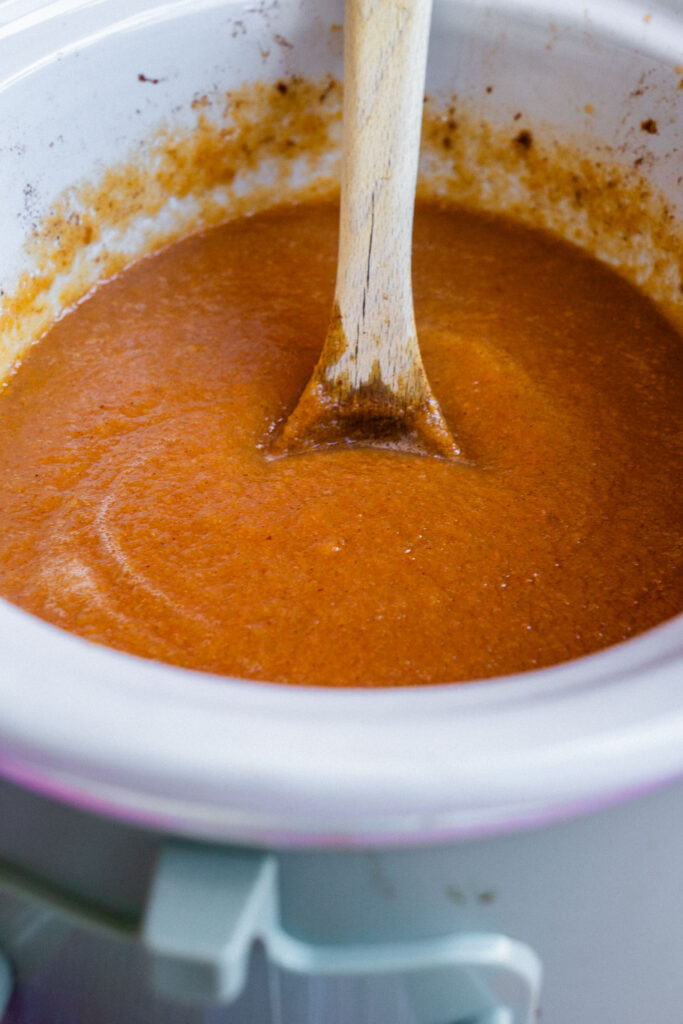 A wooden spoon stands in a slow cooker filled with a smooth pear butter.