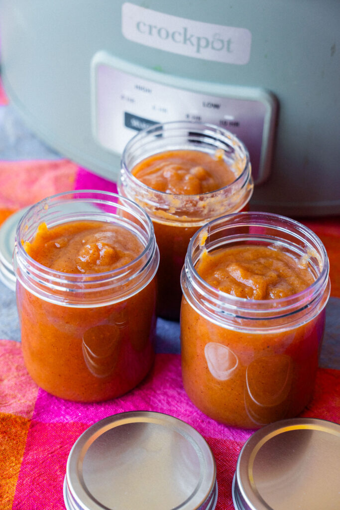 Three glass jars filled with pear butter, sitting in front of a slow cooker.